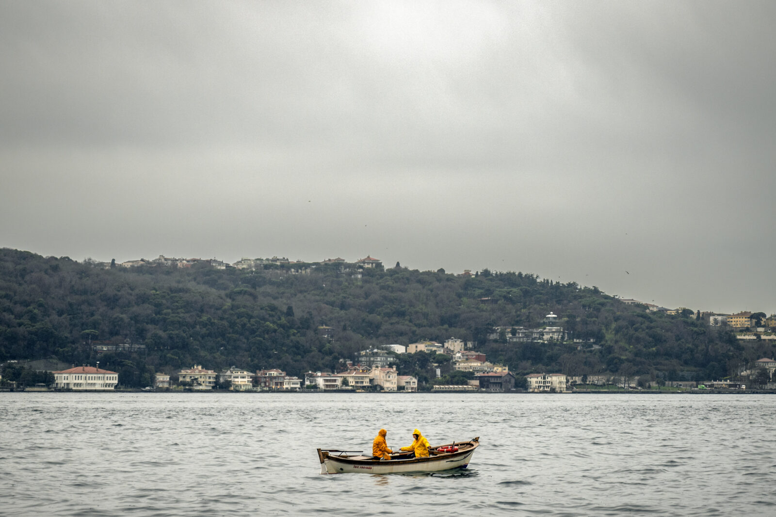 Two fishermen in bright yellow raincoats row a small wooden boat across the waters of Turkey