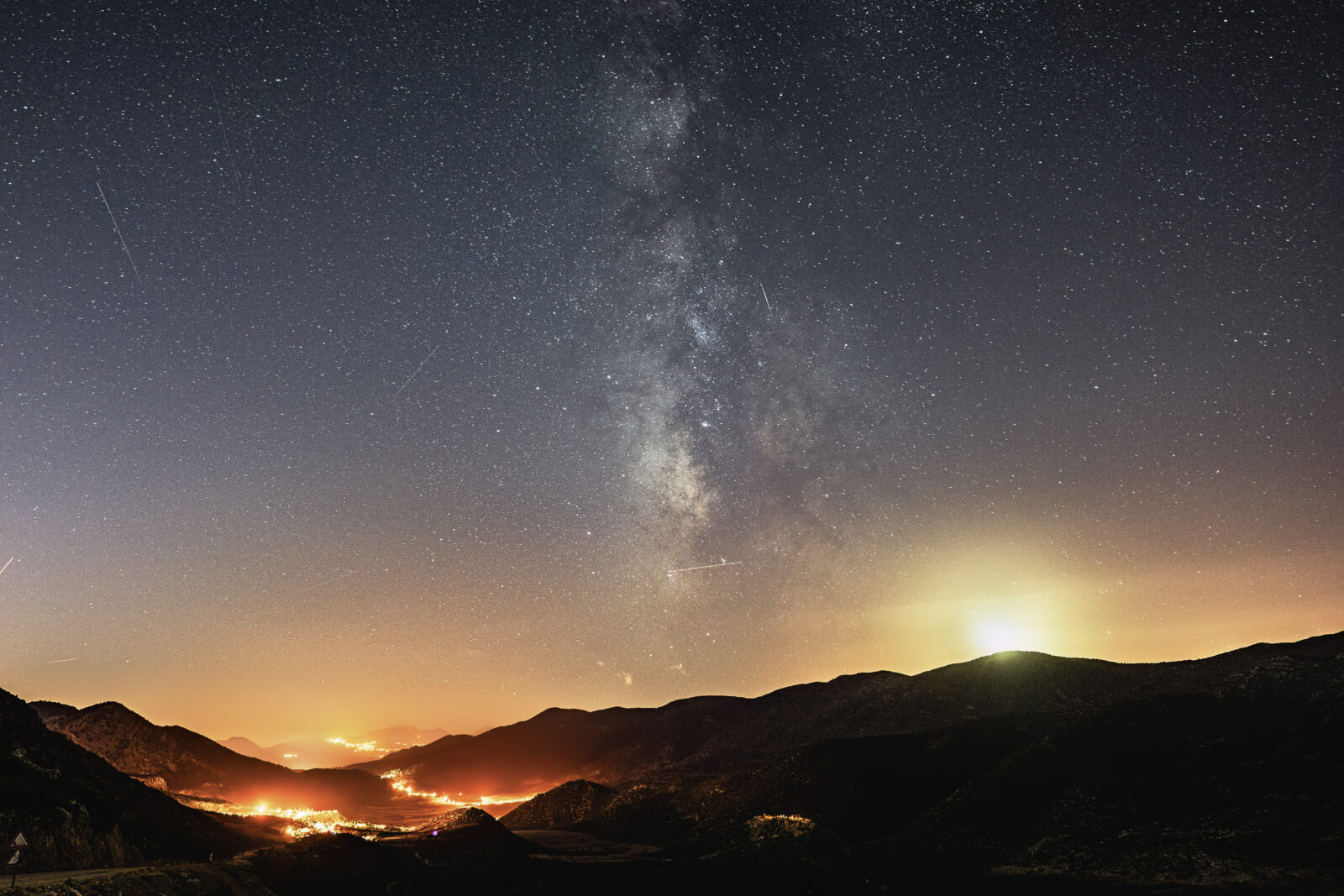 A vast star-filled sky over distant hills, illuminated by the glow of a nearby town.