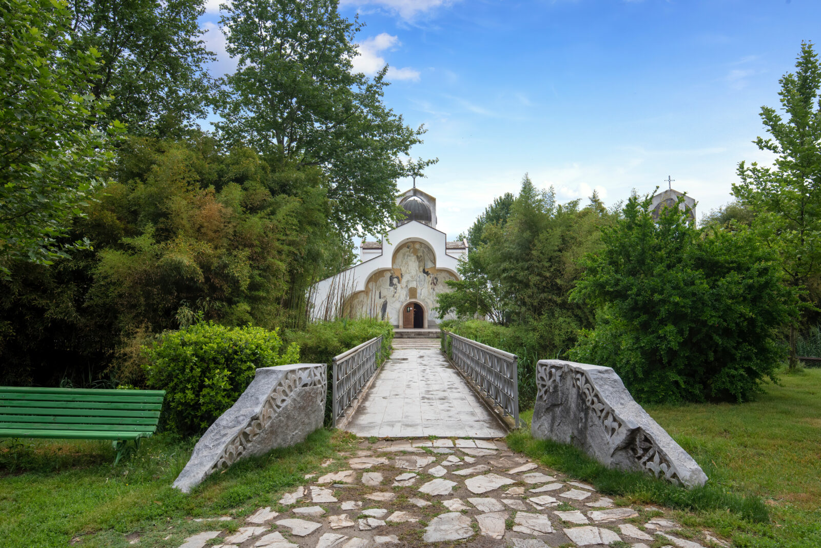 A small white Orthodox church with religious frescoes on its facade, surrounded by lush green trees and accessed via a stone bridge in a peaceful garden setting.