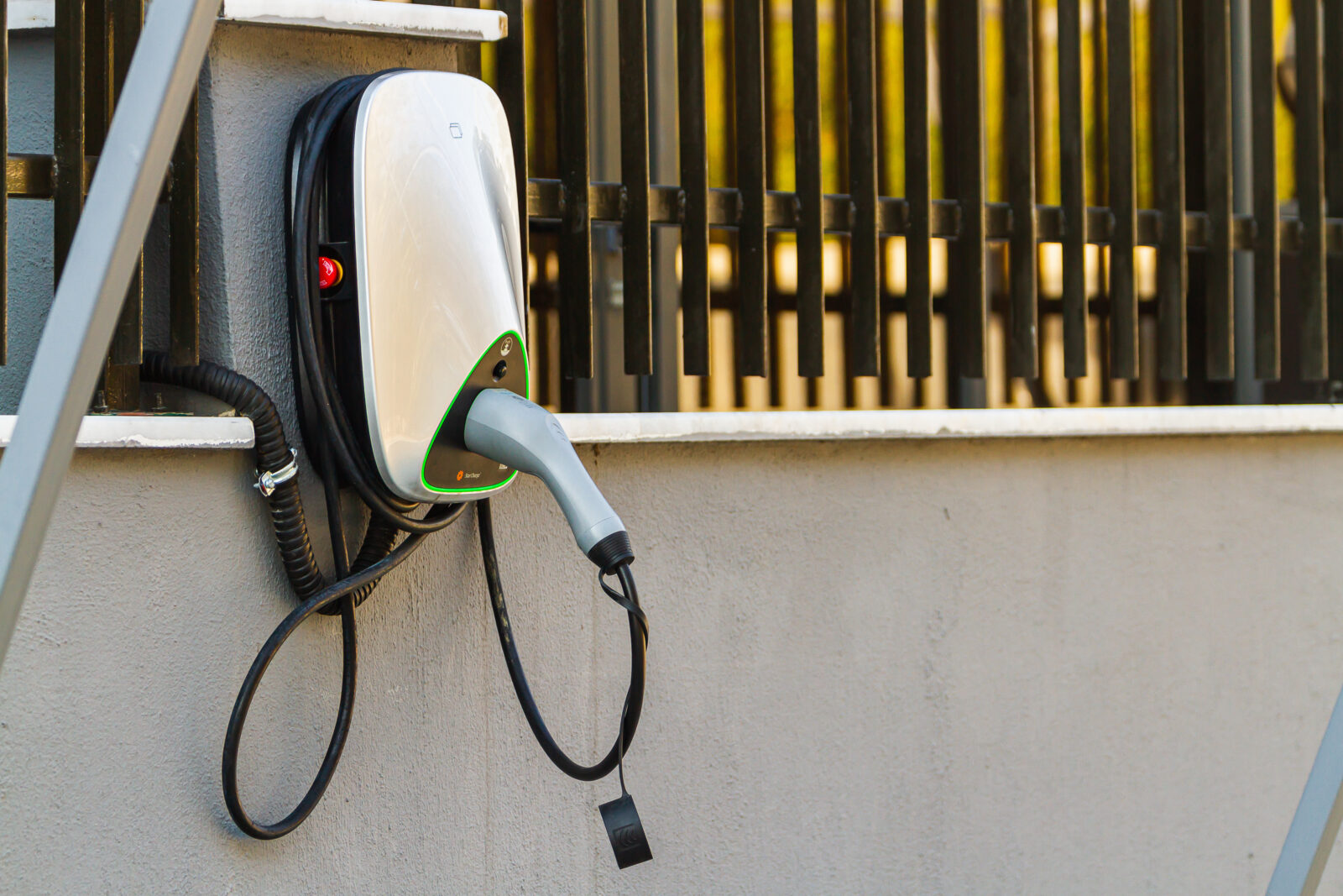 A wall-mounted EV charger attached to a concrete wall with a black fence in the background.