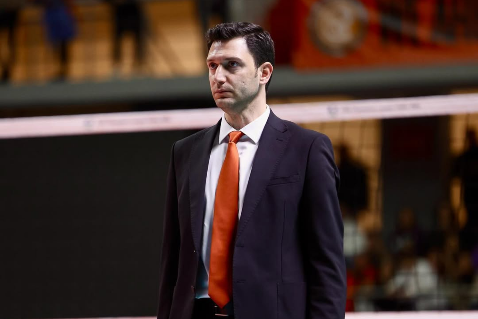 Turkish volleyball coach Ferhat Akbas standing courtside in a black suit with an orange tie, holding a tablet and appearing focused.