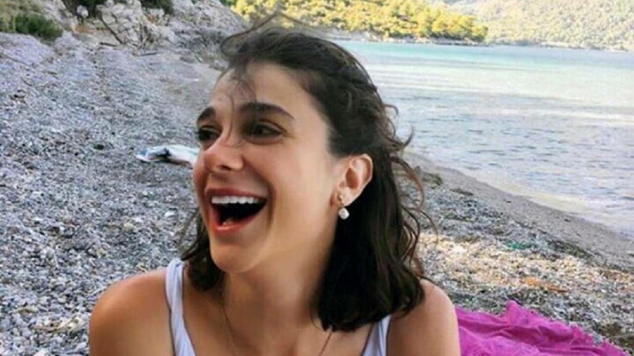 A young woman, Pinar Gultekin, smiling while sitting on a beach with the sea and greenery in the background