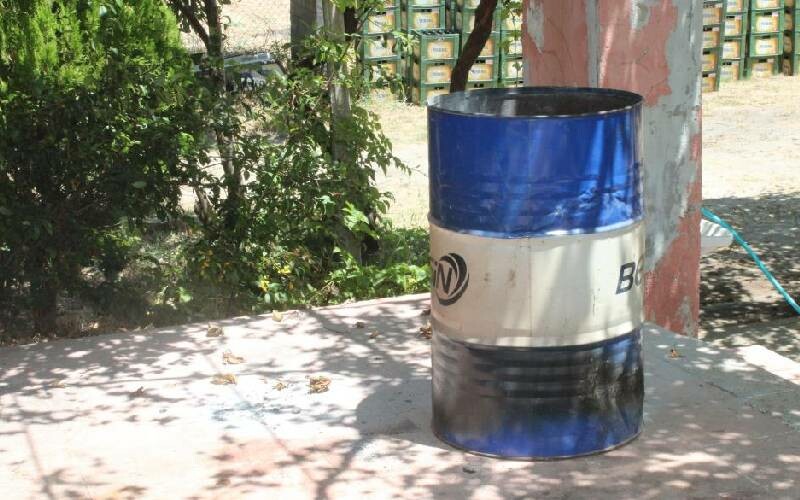 A blue and white metal barrel, partially burned, standing outdoors with trees and plants in the background