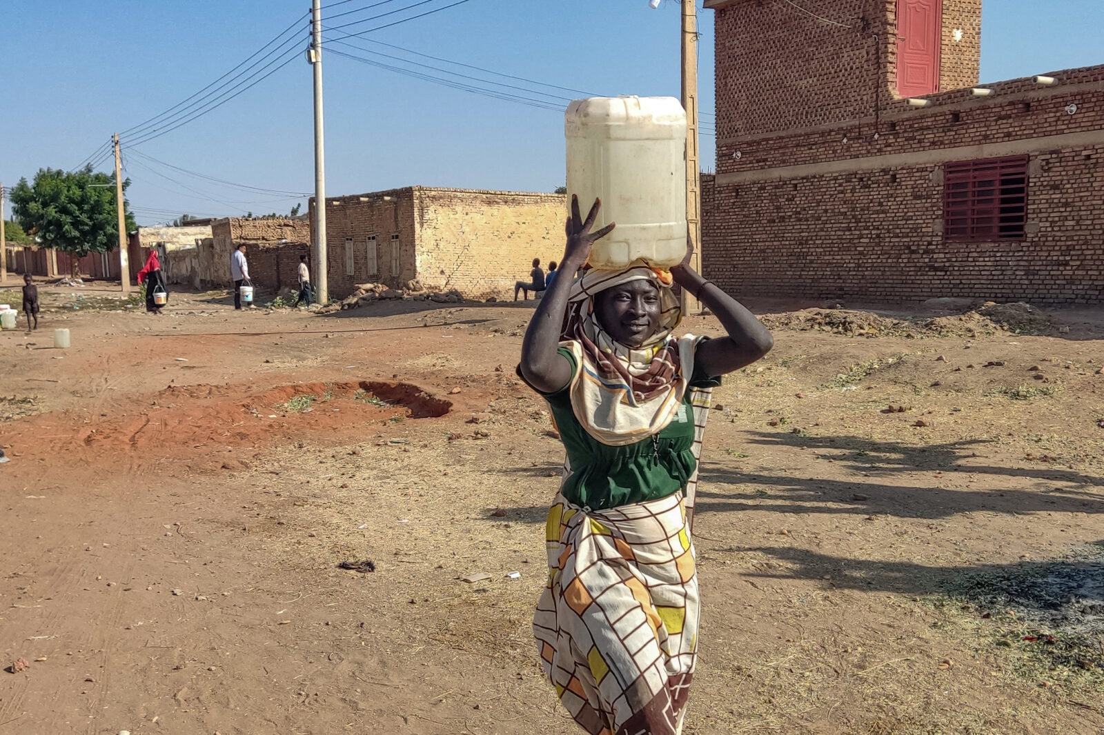 A woman smiles while carrying a large white water container on her head in a dusty neighborhood in Sudan. She wears a green top and patterned wrap skirt, standing in an unpaved street surrounded by brick buildings. In the background, other people can be seen carrying water containers, highlighting the daily water collection routine in this arid community. Power lines stretch across the clear blue sky above the settlement