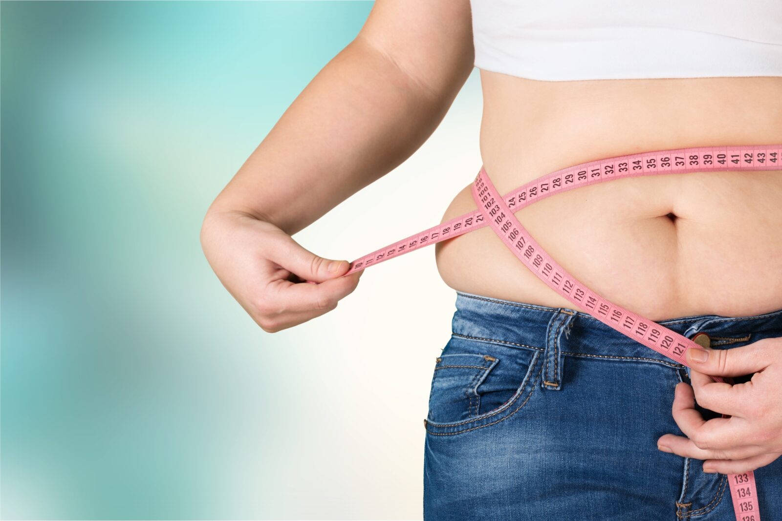 A woman in jeans and a crop top using a pink measuring tape to measure her waist, symbolizing weight control and obesity awareness.