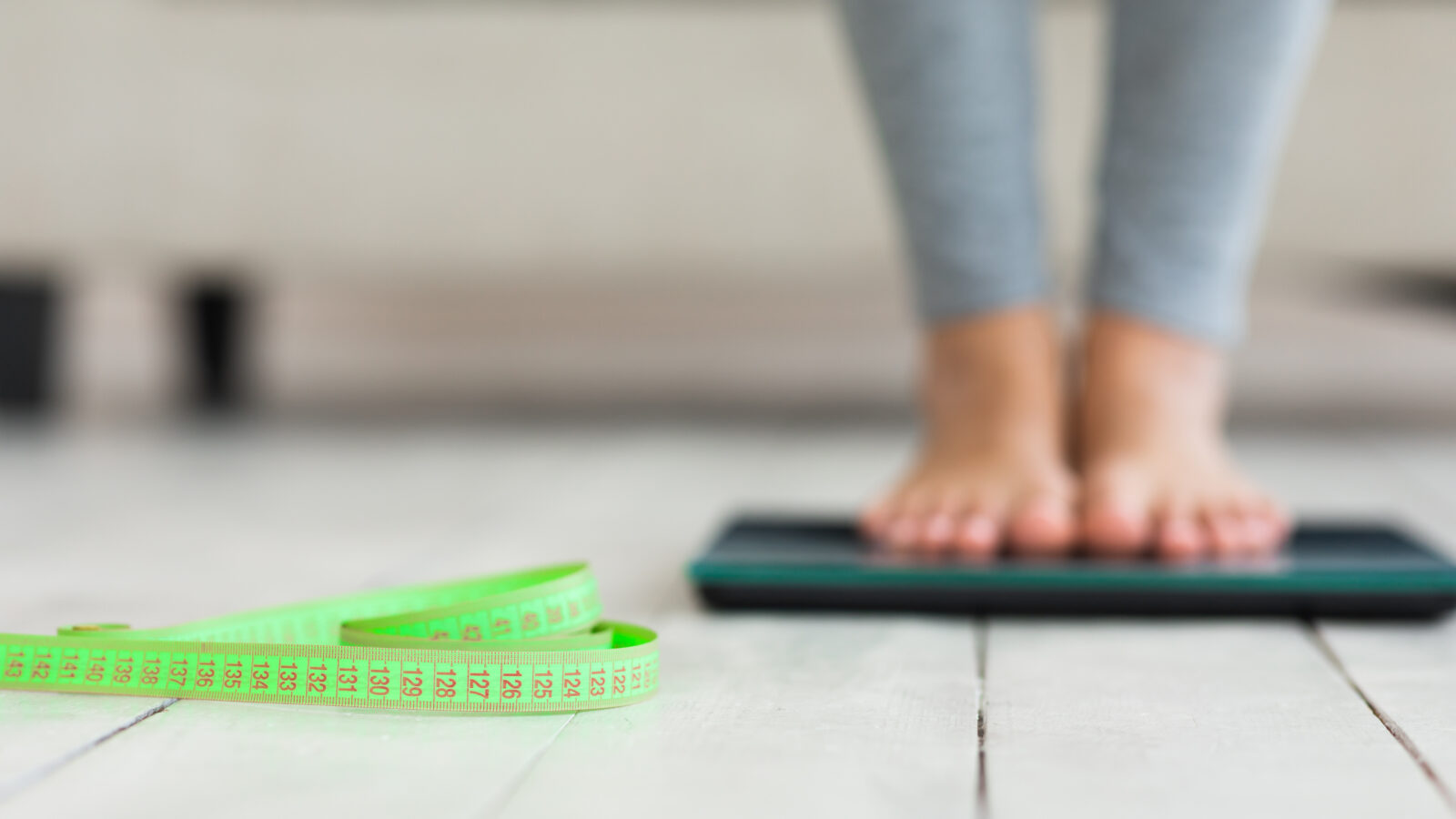 A close-up of a person’s feet on a weight scale, with a green measuring tape in the foreground, illustrating concerns about obesity and body weight tracking.
