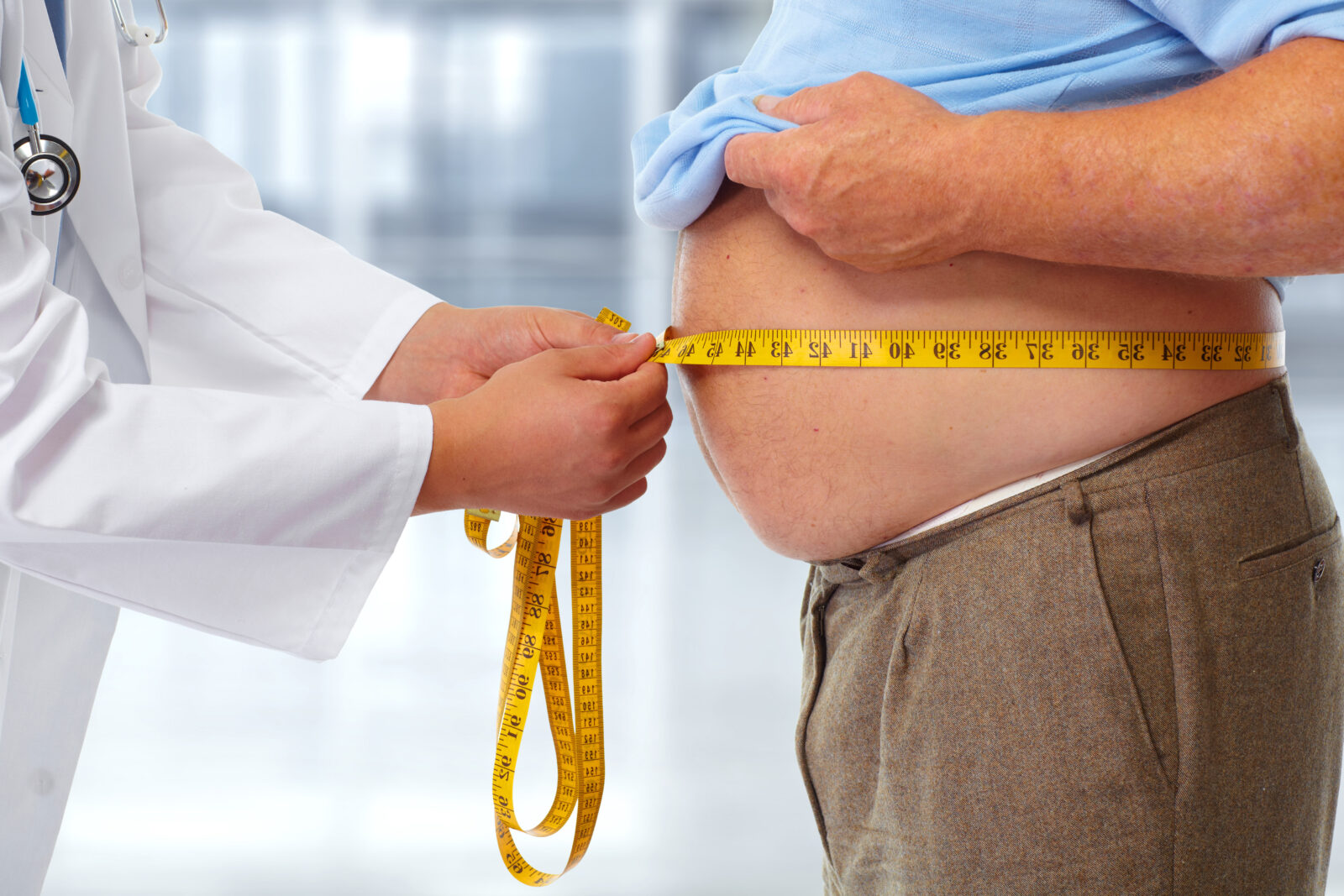 A doctor in a white coat using a yellow measuring tape to measure an overweight man