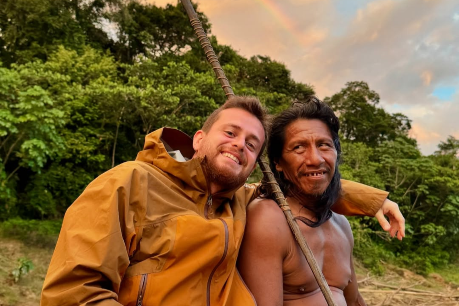 A Waorani elder with long hair, stretched earlobes, and a warm smile sits next to Alper Rende, who is wearing rain boots and making a peace sign.