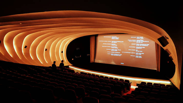 Interior of Kadikoy Cinema theater featuring a striking curved, ribbed wall design illuminated with warm amber lighting. The screen is displaying end credits of a film. Two silhouetted figures can be seen in the nearly empty theater with red seats. The architectural elements create a flowing, organic shape that wraps around the auditorium.