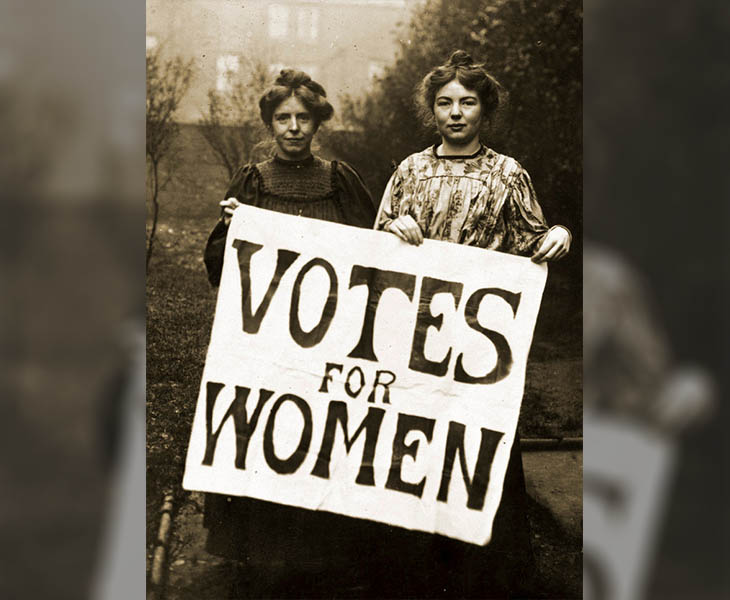 Two women from the early 20th-century suffrage movement stand holding a large ‘Votes for Women’ banner, advocating for women