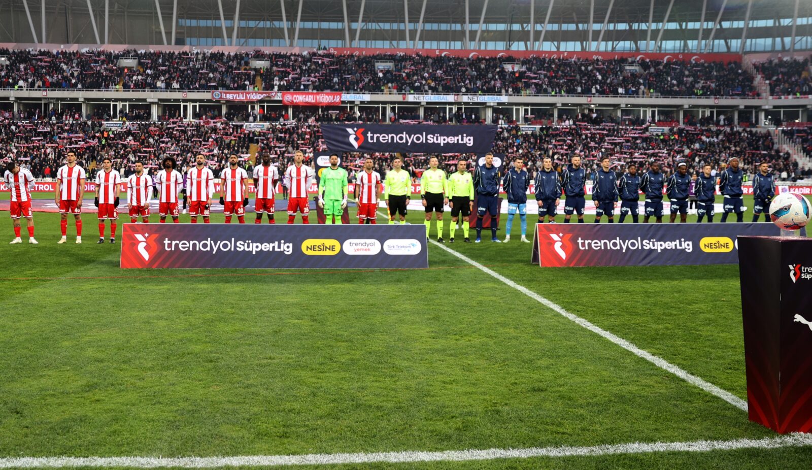 Turkish football teams Sivasspor (in red and white striped uniforms) and Fenerbahçe (in blue uniforms) lined up on the field before their match in the Trendyol Süper Lig. Referees in yellow stand between the teams. The stadium seats are filled with spectators and Trendyol Süper Lig and Nesine sponsor boards are visible at field level. This is the match where the Health Ministry