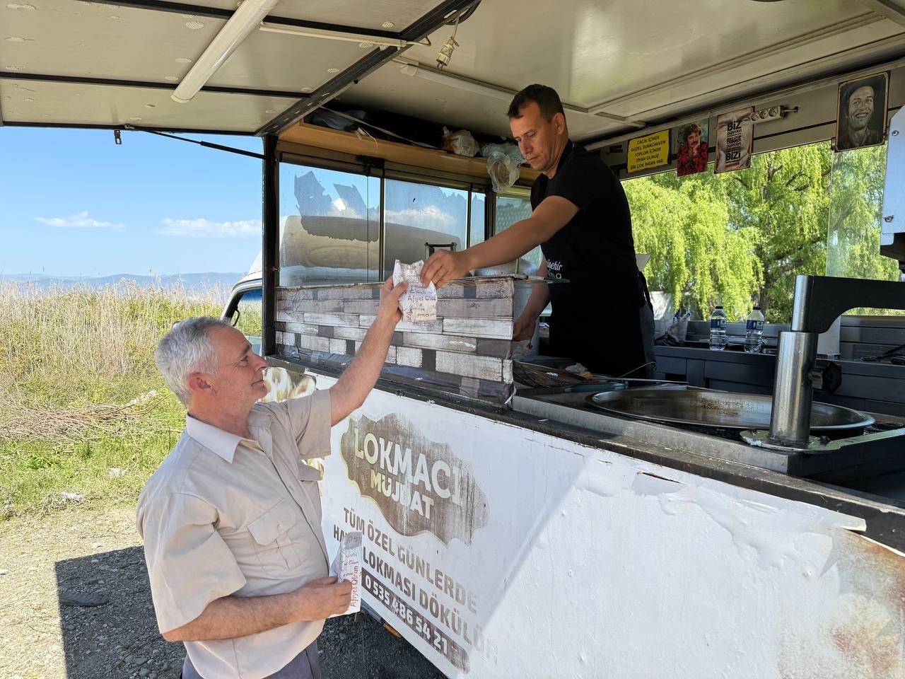 A lokma vendor in a black t-shirt stands inside his food truck handing a small package to an older man in a beige shirt standing outside. The side of the truck displays Turkish text reading 