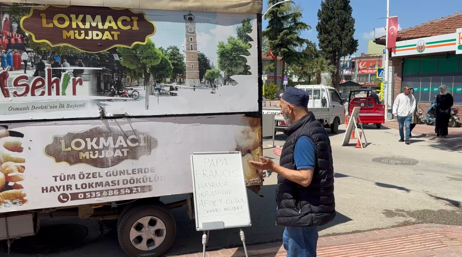 A man in a black vest and blue cap stands next to a lokma vendor