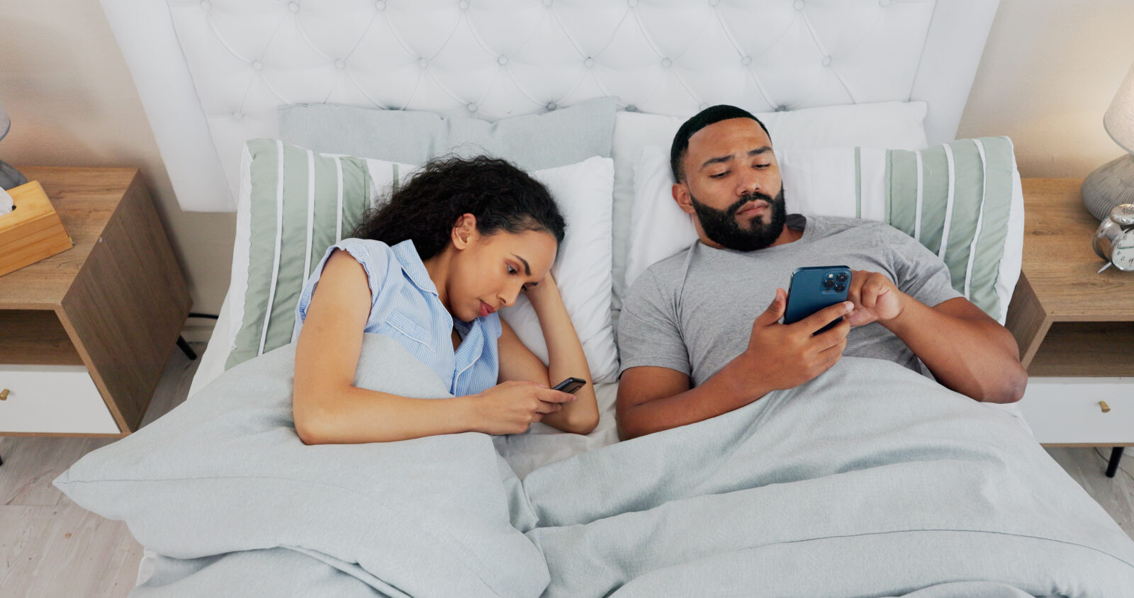 Two people in bed looking at their separate phones instead of interacting with each other, depicting smartphone usage affecting relationship intimacy.
