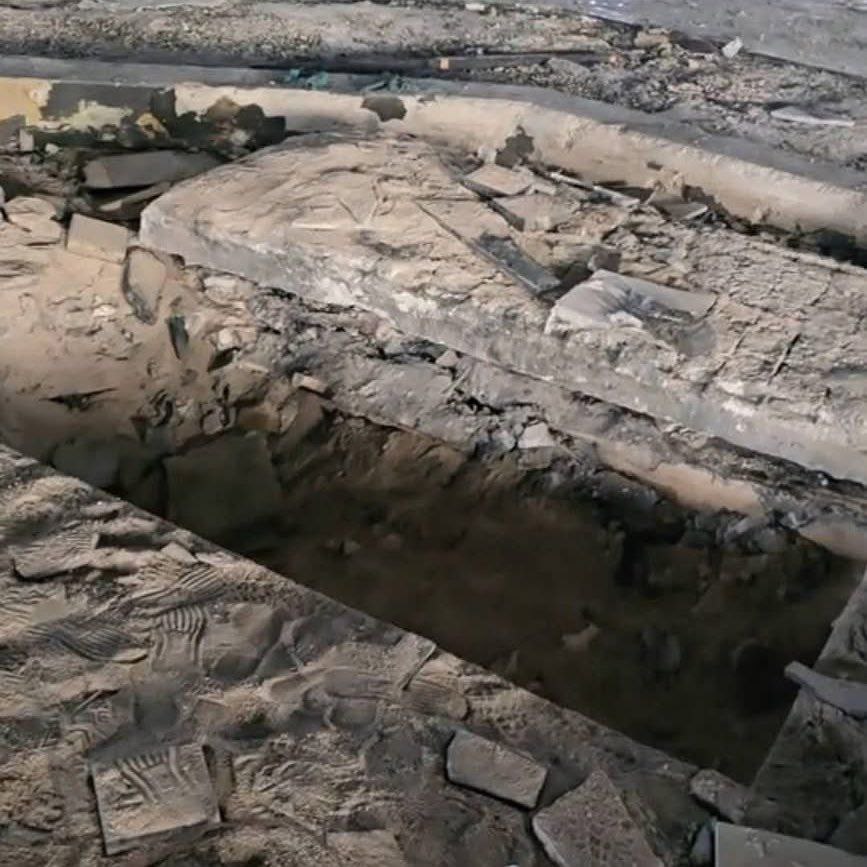 An excavated grave site with damaged stone slabs, broken concrete, and an empty burial pit with disturbed soil and rubble around the edges